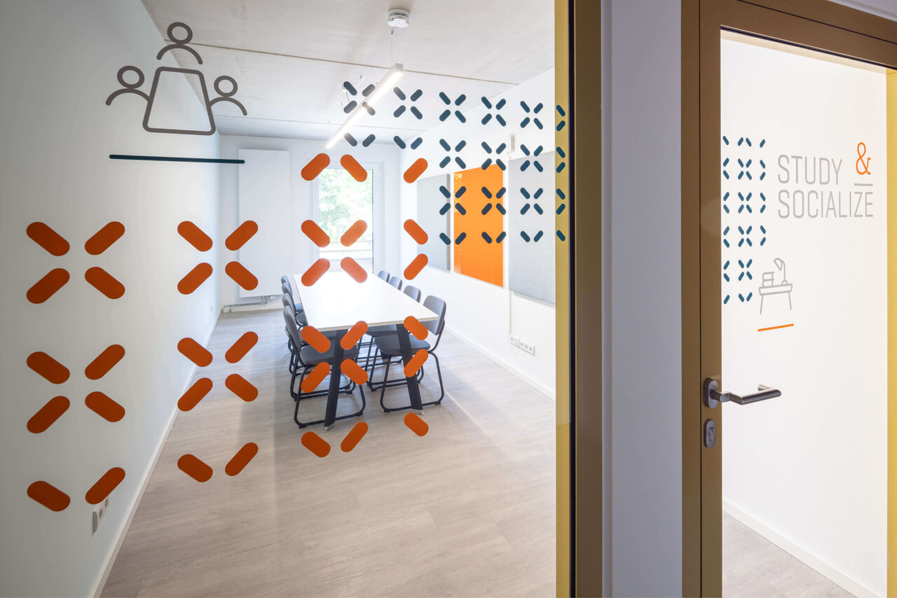 Moderner Besprechungsraum mit grafischen orange-wei&szlig;en Wandelementen, grauen St&uuml;hlen um einen Tisch und einem T&uuml;rschild mit "Study & Socialize" in minimalistischem Design.