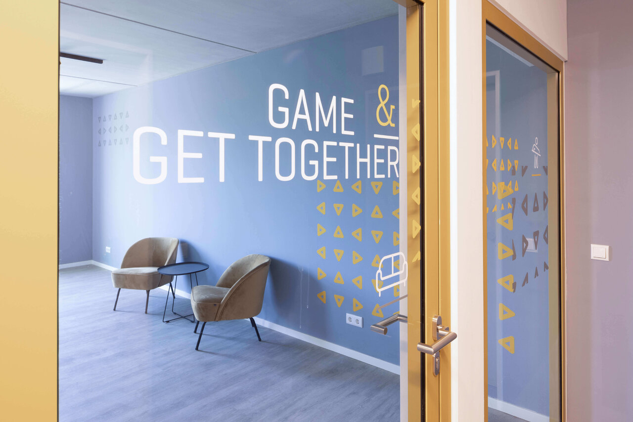 Moderner Meetingraum mit blauer Wand und gelber Beschriftung "GET TOGETHER & GAME", ausgestattet mit zwei braunen Sesseln und Holzt&uuml;r mit zus&auml;tzlichen Wandgrafiken.