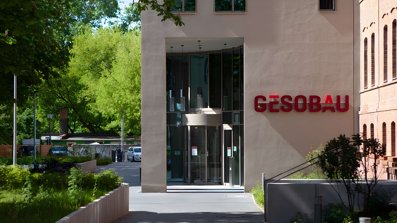 Eingang des GESOBAU-Geb&auml;udes mit moderner Glasfront und rotem Firmenlogo, umgeben von gr&uuml;ner Bepflanzung und einem gepflegten Vorplatz in urbaner Umgebung.