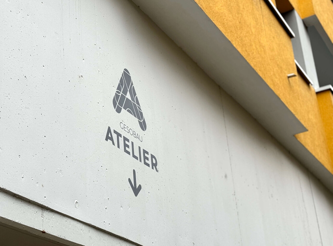 Wei&szlig;e Wand mit grauem ATELIER-Logo und Pfeil, daneben eine gelbe Wand, Teil eines Geb&auml;udes oder Eingangsbereichs.