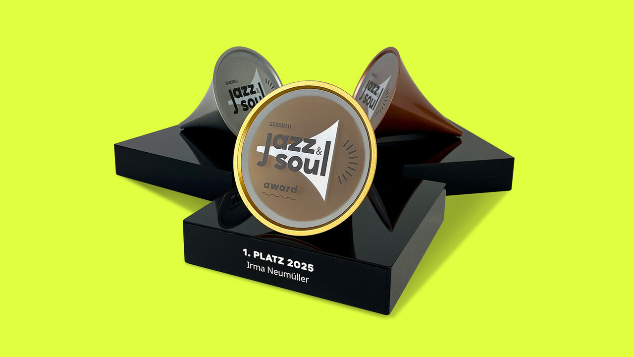Ein Award in Form dreier Metalltrichter (Gold, Graphit, Kupfer) auf schwarzem Stufensockel. Vorderer Trichter tr&auml;gt &bdquo;GESOBAU jazz & soul award&ldquo;, der Sockel &bdquo;1. PLATZ 2025 &ndash; Irma Neum&uuml;ller&ldquo;. Hintergrund: leuchtendes Neon-Gelb.