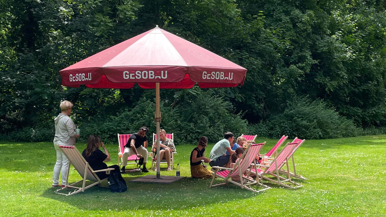 Mehrere Personen entspannen auf Liegest&uuml;hlen unter einem roten GESOBAU-Sonnenschirm auf einer gr&uuml;nen Wiese mit B&auml;umen im Hintergrund.