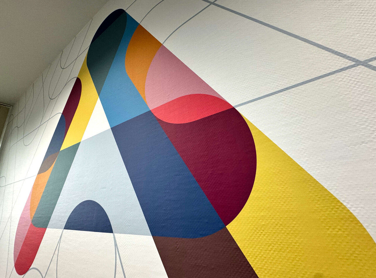 Abstrakte Wandgestaltung mit &uuml;berlappenden geometrischen Formen in kr&auml;ftigen Farben wie Gelb, Blau, Rot und Rosa, die eine dynamische Komposition auf einer wei&szlig;en Wand bilden.