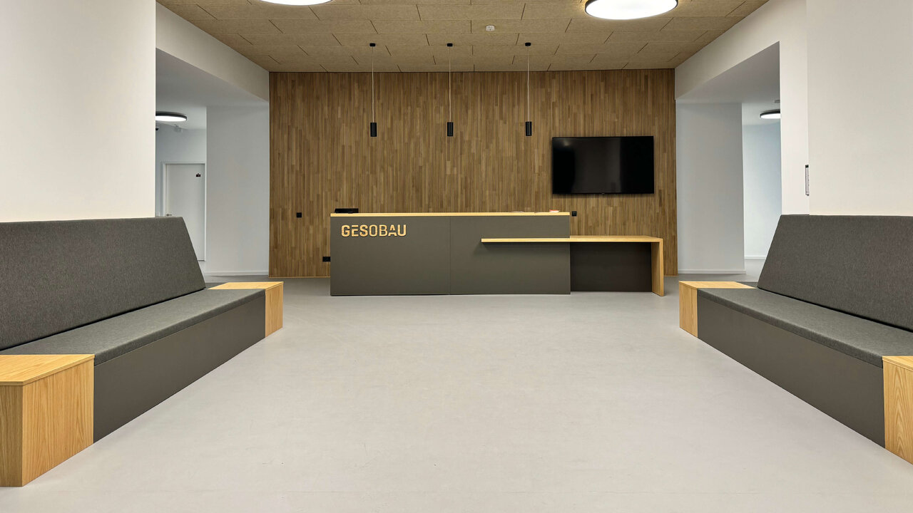 Empfangsbereich eines Workspaces der GESOBAU mit minimalistischem Design, grauen Sitzgelegenheiten und Holzelementen, einer Rezeption mit Firmenlogo und modernen Deckenleuchten in einem hellen, geradlinigen Raum.