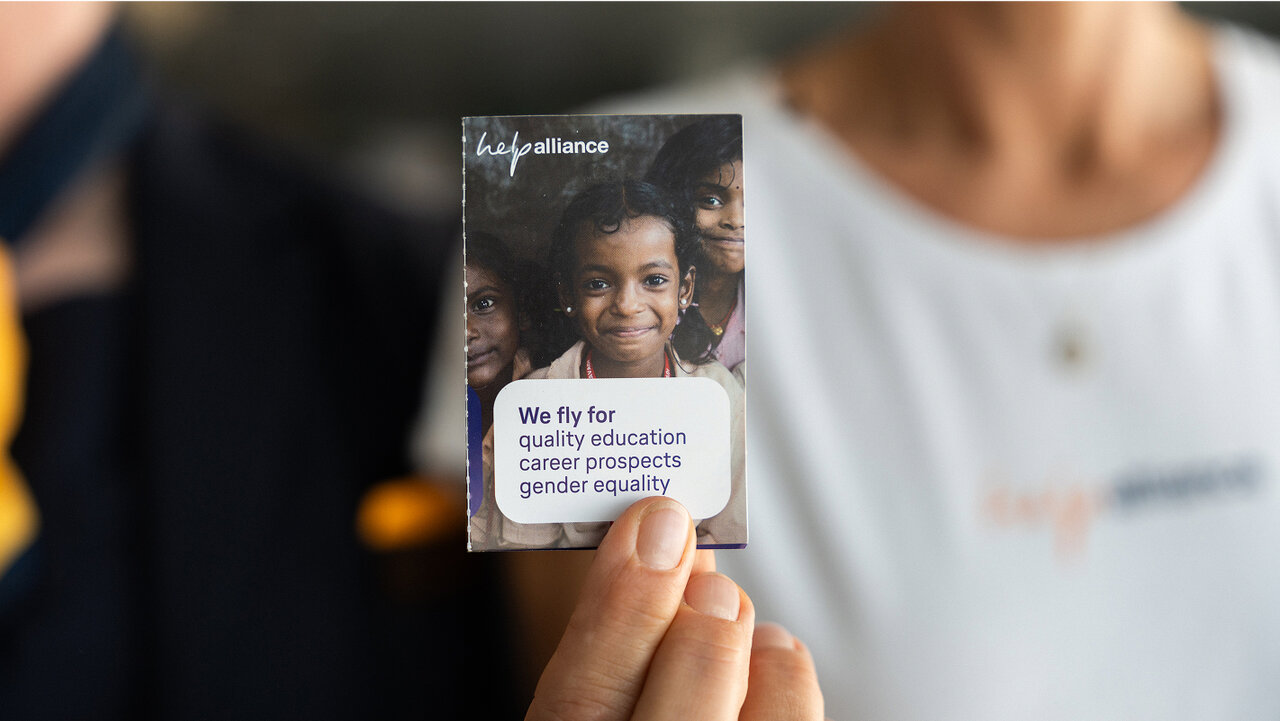 Eine Hand h&auml;lt eine Visitenkarte der Kids Alliance mit einem Bild l&auml;chelnder Kinder und dem Text "We fly for quality education, career prospects, gender equality".