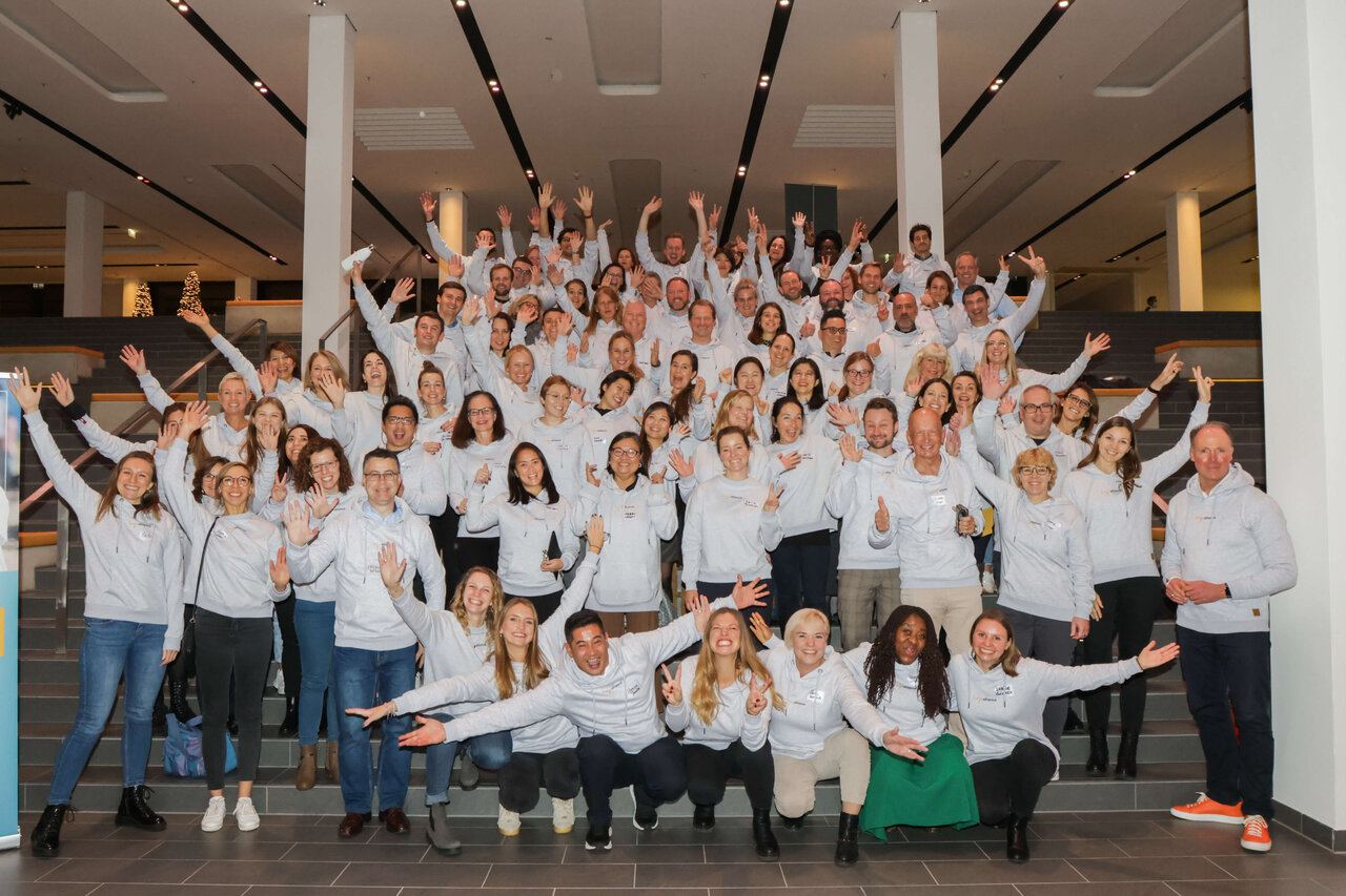 Gruppenaufnahme des Studio Good Teams in wei&szlig;en Oberteilen, die gemeinsam l&auml;cheln und jubeln. Etwa 50 Mitarbeiter posieren auf Treppenstufen in einem hellen Innenraum, viele mit erhobenen Armen in einer fr&ouml;hlichen, enthusiastischen Haltung.