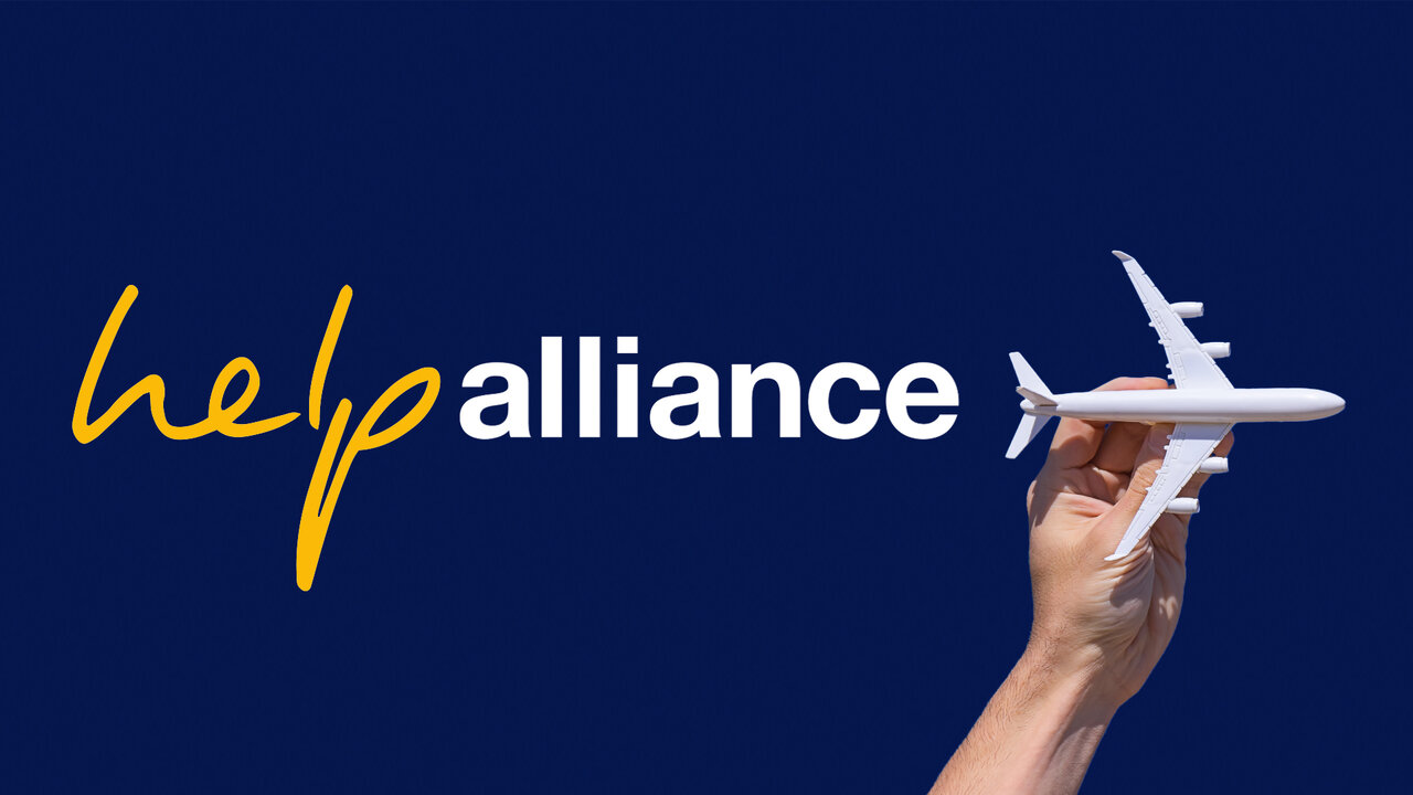 Hand hebt ein wei&szlig;es Flugzeugmodell vor blauem Hintergrund mit dem Logo von "Help Alliance" in gelber und blauer Schrift.
