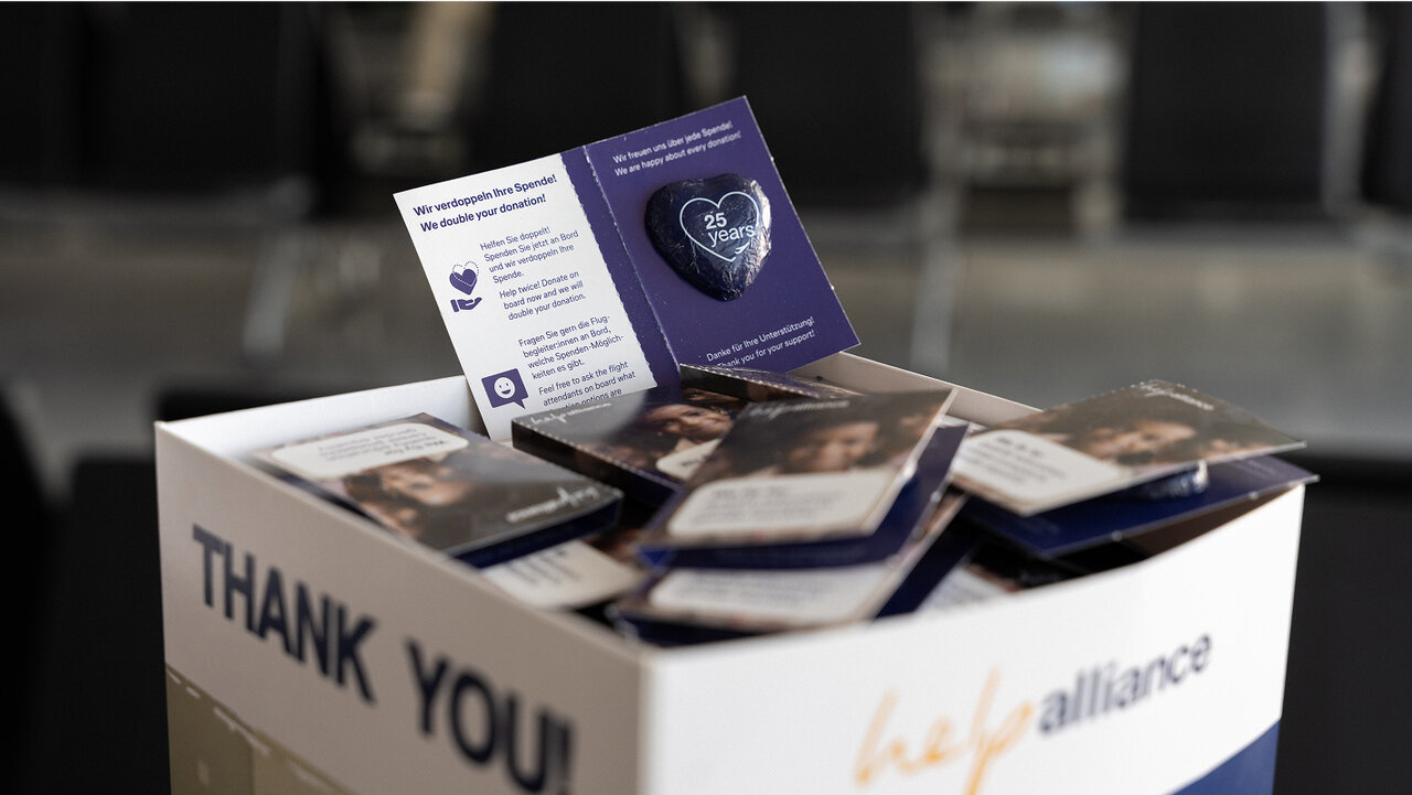 Eine Kartonschachtel mit "THANK YOU" Aufdruck, gef&uuml;llt mit lila und blauen Brosch&uuml;ren und Informationsmaterialien von Alliance, pr&auml;sentiert auf einem unscharfen Hintergrund.