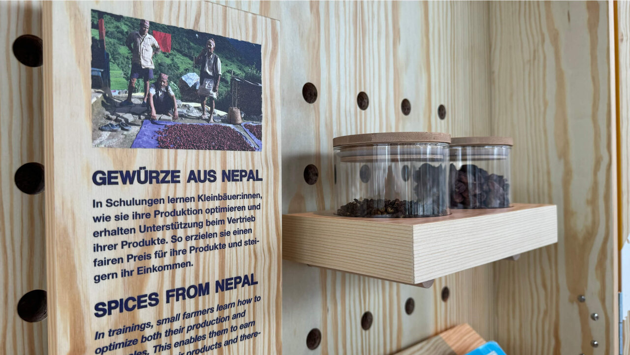 Holzregal mit Gew&uuml;rzgl&auml;sern und einem Informationsplakat &uuml;ber Gew&uuml;rze aus Nepal, zeigt eine Nahaufnahme der Ausstellung mit Gew&uuml;rzen in Gl&auml;sern vor einer Holzwand mit L&ouml;chern.