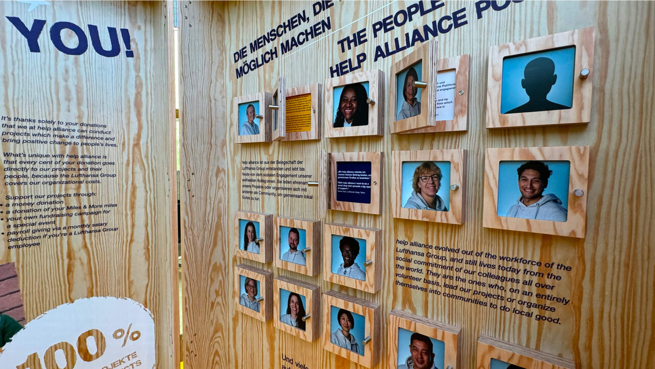 Holzwand mit Fotowand zeigt Portr&auml;ts verschiedener Menschen in blau-grauen T&ouml;nen, Teil einer Ausstellung &uuml;ber gesellschaftliches Engagement und Gemeinschaftsprojekt, mit Texten auf Deutsch und Englisch &uuml;ber Zusammenarbeit und Hilfsallianzen.