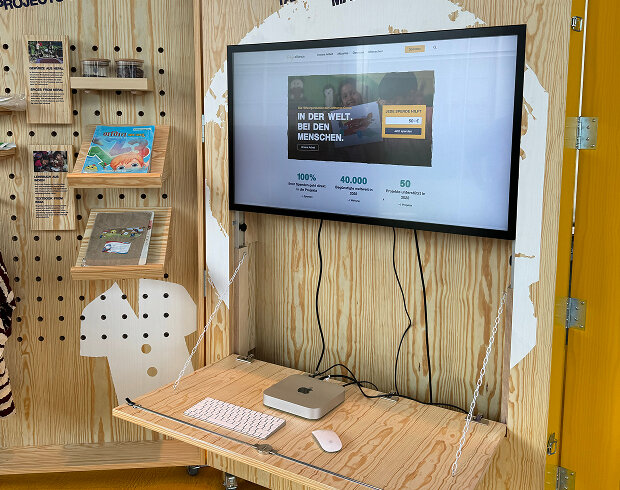 Ein Arbeitsplatz mit einem Bildschirm, der eine Website zeigt, auf einem h&ouml;lzernen Regal mit Aufh&auml;ngungen, darunter ein Macbook und eine Tastatur, in einem kreativ gestalteten B&uuml;robereich von Studio Good in Berlin.