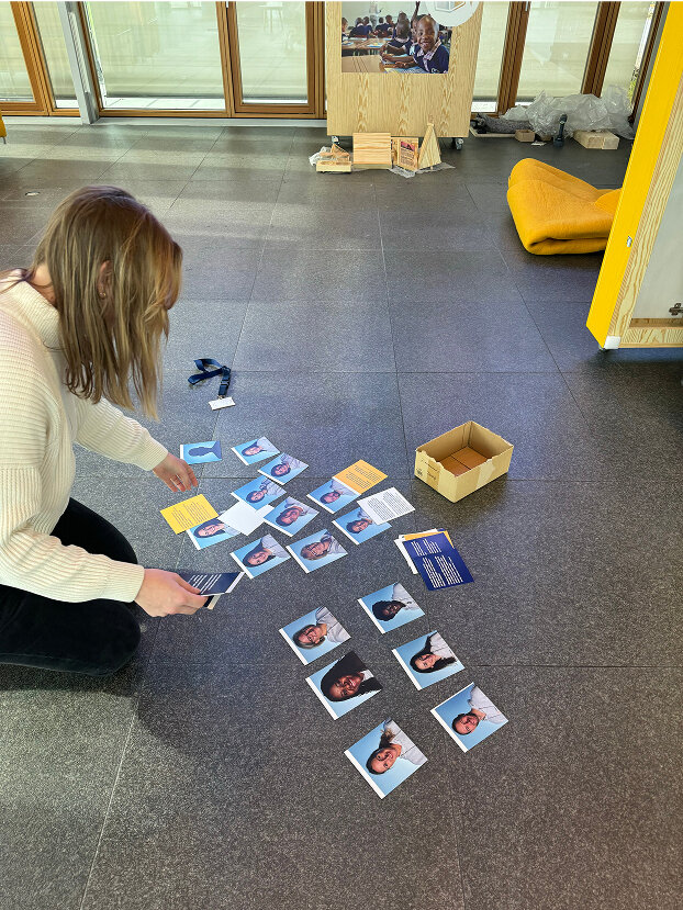 Eine Person ordnet Polaroid-&auml;hnliche Fotos auf dem Boden an, w&auml;hrend sie an einem Memory-Spiel zu arbeiten scheint. Die Fotos zeigen Portraitaufnahmen, und eine Holzkiste steht daneben auf dem grauen Teppichboden.