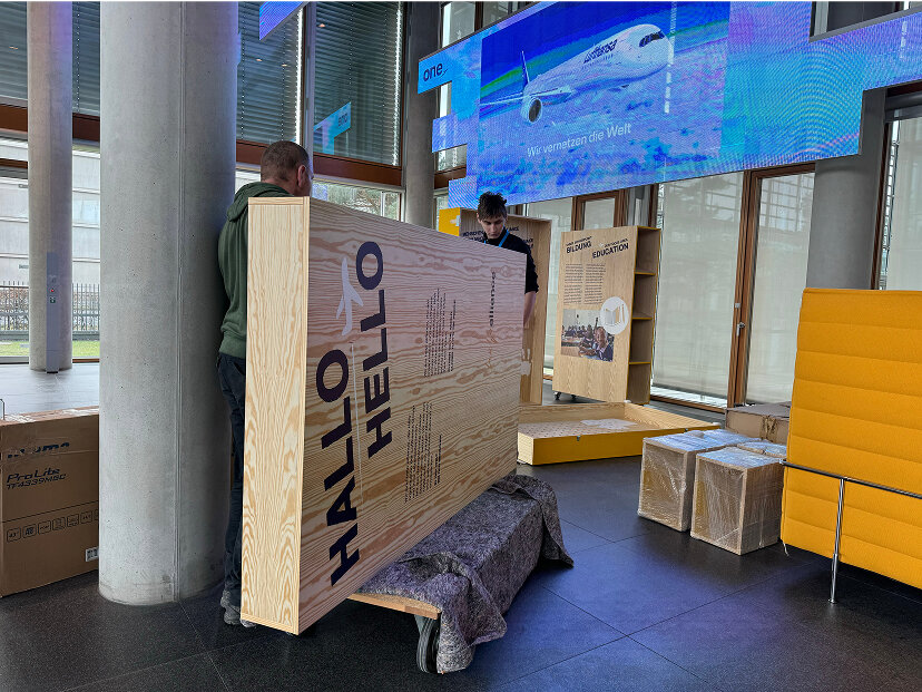 H&ouml;lzerne Ausstellungstafel mit "HALLO" Beschriftung in einem modernen B&uuml;roraum mit gelben Sitzm&ouml;beln und gro&szlig;er blauer Bildschirmanzeige im Hintergrund.