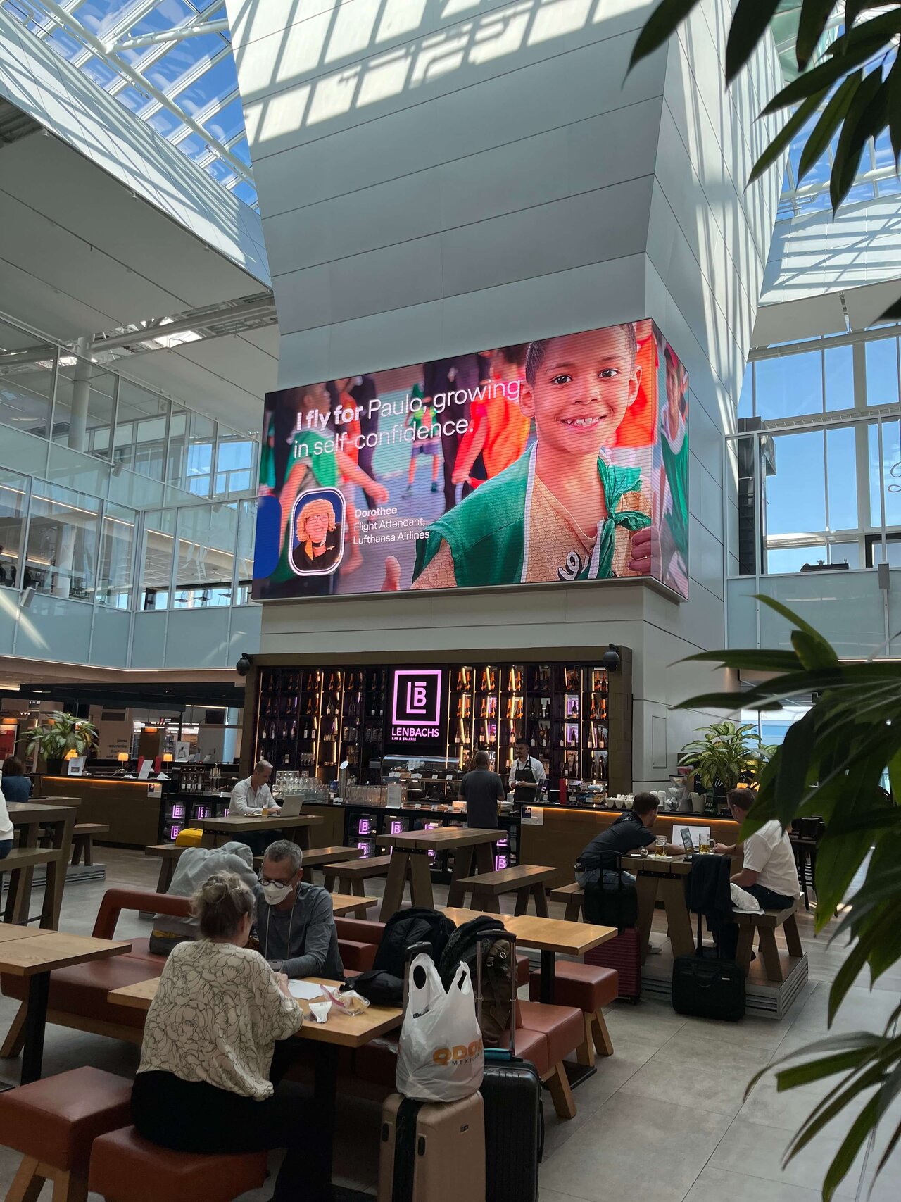 Gro&szlig;er Innenraum mit Caf&eacute; und &uuml;bergro&szlig;er LED-Wand, die ein Werbemotiv mit einem l&auml;chelnden Kind in gr&uuml;ner Kleidung zeigt. Holzm&ouml;bel und Pflanzen pr&auml;gen die moderne, lichtdurchflutete Architektur mit Glasdach.