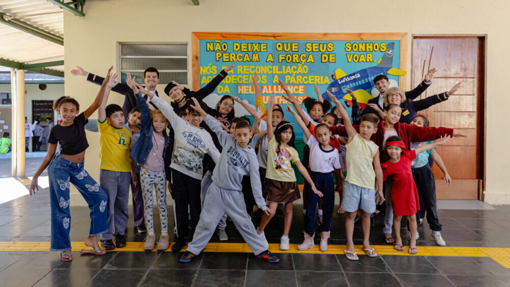 Eine lebhafte Gruppe von Kindern posiert zusammen mit ausgestreckten Armen und dynamischen Posen vor einem bunten Wandbild in einem Schulflur. Die Kinder tragen verschiedene Kleidungsst&uuml;cke und strahlen Freude und Energie aus. Sie scheinen Teil einer Schulgemeinschaft zu sein, die Spa&szlig; und Zusammenhalt zelebriert.