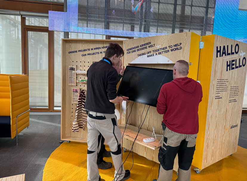 Zwei Personen stehen an einem Holzstand mit interaktiver Technik, umgeben von einer gelben Bodenfl&auml;che und Glasw&auml;nden in einem modernen Ausstellungsraum.