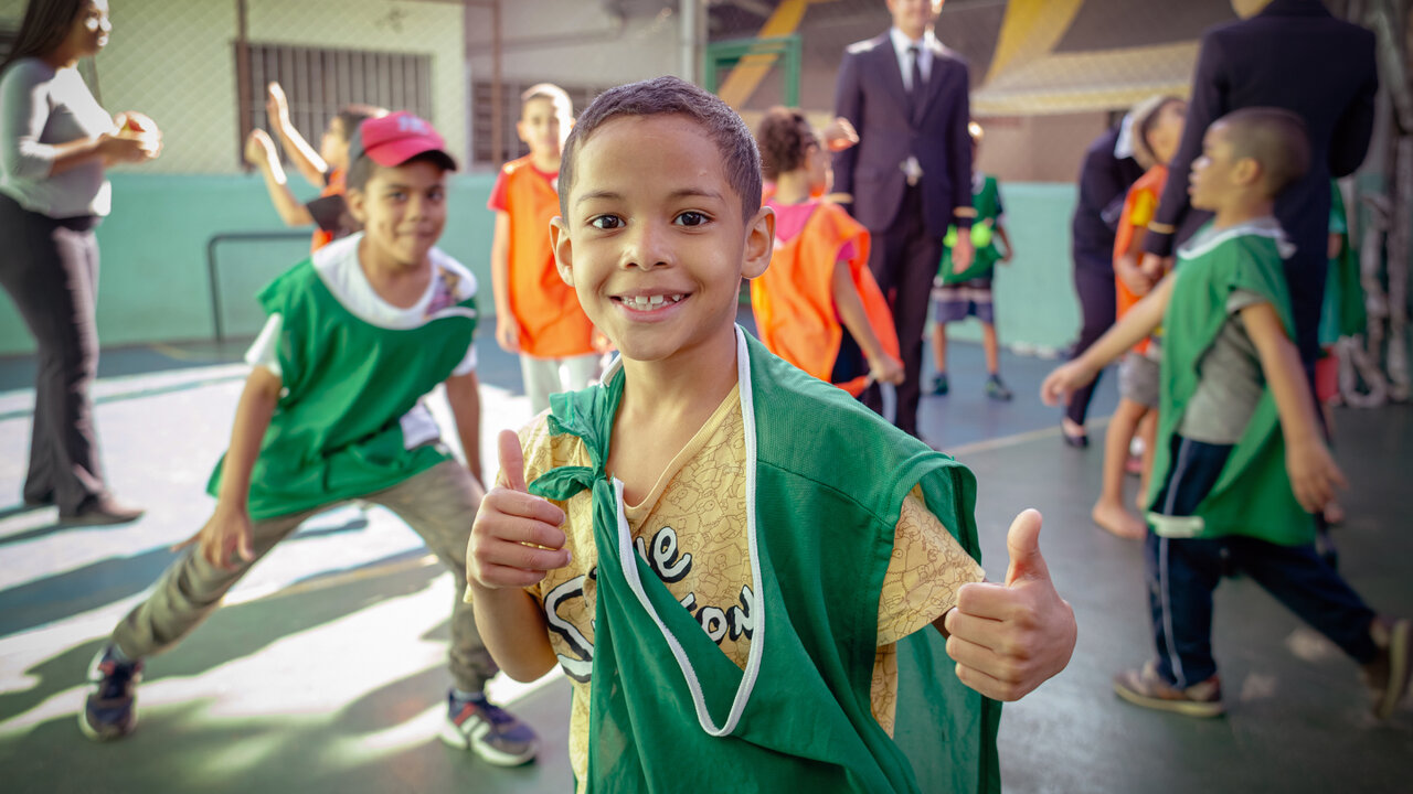 Ein l&auml;chelnder Junge in gr&uuml;nem Sportdress gibt Daumen hoch, umgeben von anderen Kindern in einem Schulflur oder Sportzentrum. Seine K&ouml;rpersprache strahlt Freude und Enthusiasmus aus.