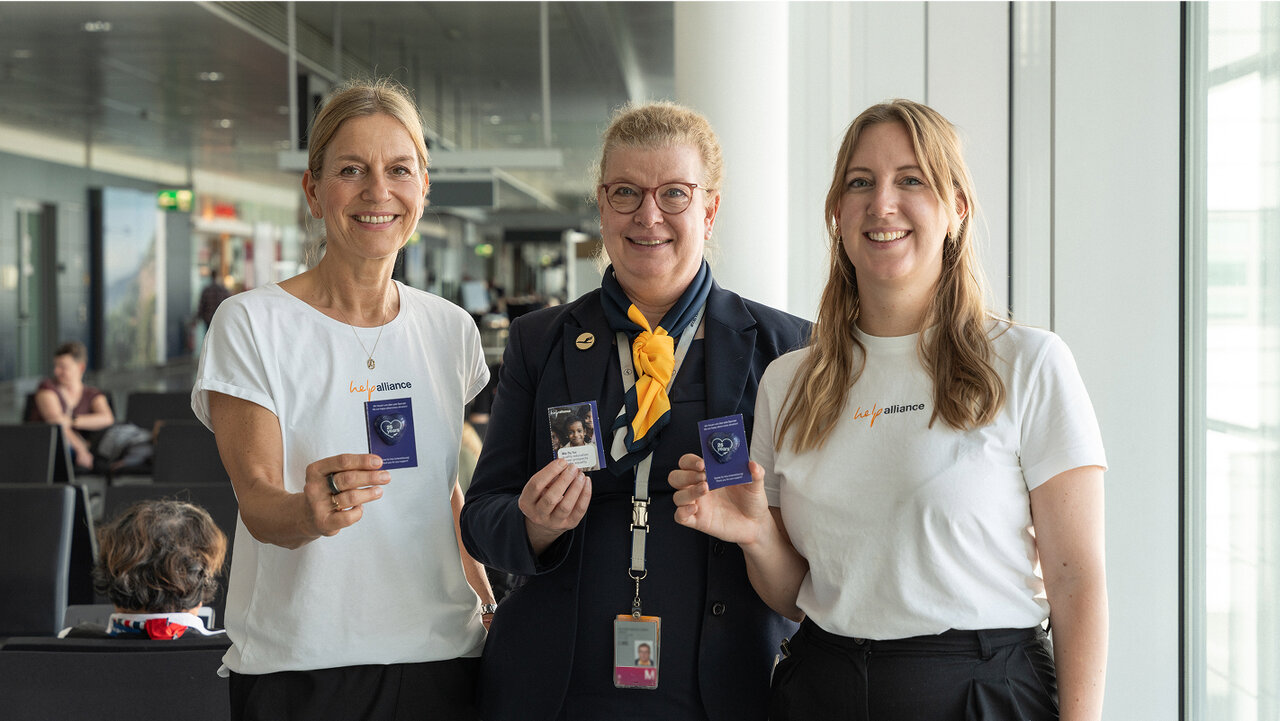 Drei Mitarbeiterinnen eines Unternehmens stehen l&auml;chelnd in einem B&uuml;rogeb&auml;ude und halten Ausweise oder Badges in den H&auml;nden. Sie tragen wei&szlig;e T-Shirts und eine Frau tr&auml;gt zus&auml;tzlich eine dunkle Jacke mit einem Firmen-Lanyard.