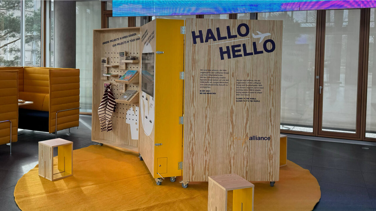 H&ouml;lzerne Ausstellungswand mit gelbem Akzent und der Aufschrift "HALLO HELLO" in einem modernen B&uuml;roraum mit Holzboden und Glasfenstern. Mehrere Holzhocker und ein gelber Raumteiler mit verschiedenen Informationstafeln bilden die Szenerie.