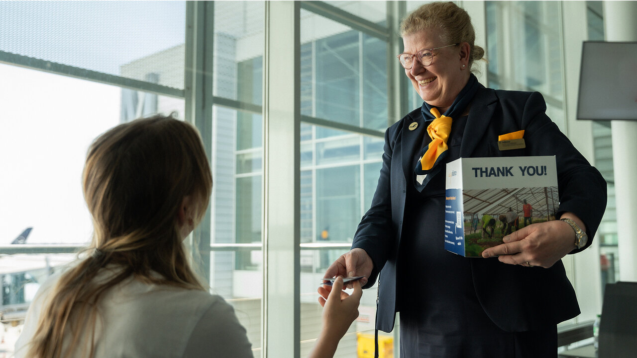 Eine freundliche Mitarbeiterin mit gelber Schleife l&auml;chelt und h&auml;lt ein "Danke sch&ouml;n"-Schild in einem hellen, modernen Flughafenbereich beim Interagieren mit einer Kundin.