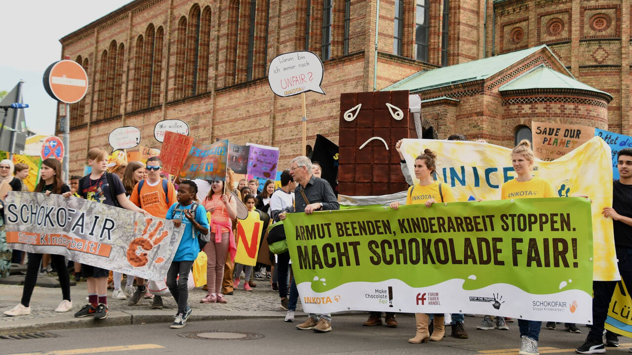 Demonstration gegen Kinderarbeit mit Plakaten und Menschen, die faire Schokolade fordern. Protest vor einem historischen Backsteingeb&auml;ude mit gelben und bunten Transparenten, die eine faire Schokoladenproduktion verlangen.