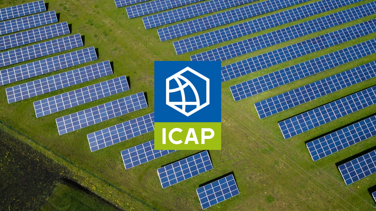 Luftaufnahme eines Solarparks mit blauen Photovoltaik-Modulen auf gr&uuml;ner Wiese, in der Mitte das ICAP-Logo auf blauem Hintergrund. Die Solarpanels sind in Reihen angeordnet und zeigen die Ausrichtung auf nachhaltige Energieerzeugung.