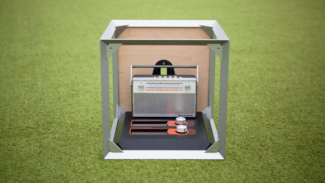 Mobiles Ausstellungsmodul mit integriertem Bildschirm und zwei orangefarbenen Elementen, eingebettet in einen minimalistischen Metallrahmen auf gr&uuml;nem Hintergrund, entwickelt f&uuml;r die on.tour Ausstellung des J&uuml;dischen Museums Berlin.