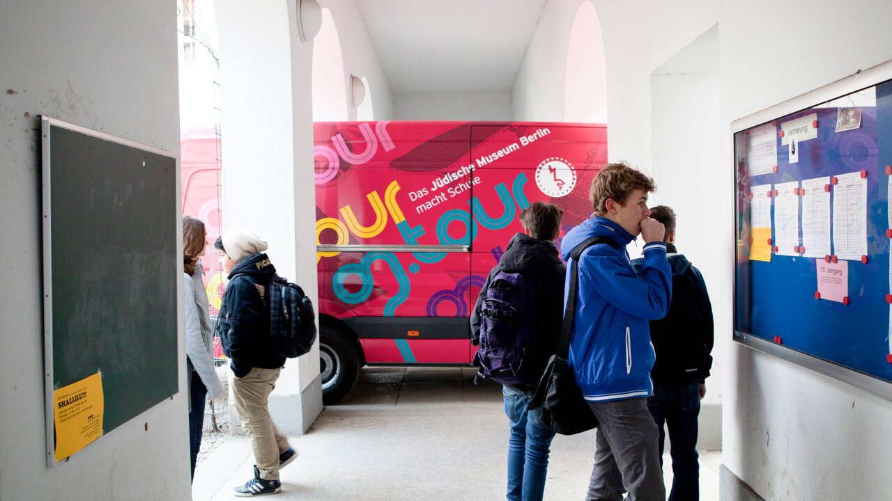 Sch&uuml;ler im Flur einer Schule stehen vor einem bunten rot-pinken Ausstellungsplakat, das Teil der mobilen on.tour Ausstellung des J&uuml;dischen Museums Berlin ist. Verschiedene Personen tragen Jacken und Rucks&auml;cke und betrachten Informationstafeln an den W&auml;nden.