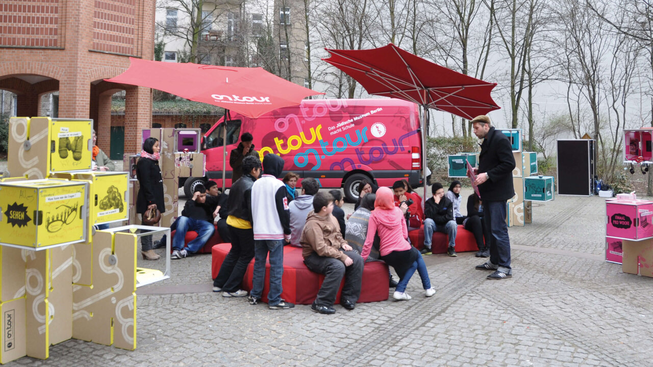 Eine mobile Ausstellung des J&uuml;dischen Museums Berlin mit einem pinkfarbenen Truck, roten Sonnenschirmen und interaktiven Ausstellungsmodulen auf einem Platz, umgeben von Besuchern und Sch&uuml;lern, die an den St&auml;nden interagieren.