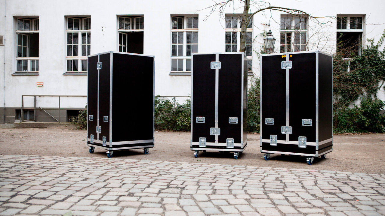 Drei schwarze transportable Ausstellungsmodule auf Rollen stehen auf einem Kopfsteinpflaster vor einem wei&szlig;en Geb&auml;ude. Die robusten Transportkoffer sind ger&auml;umig und mit Metallbeschl&auml;gen versehen, bereit f&uuml;r den flexiblen Einsatz der mobilen Ausstellung des J&uuml;dischen Museums Berlin.