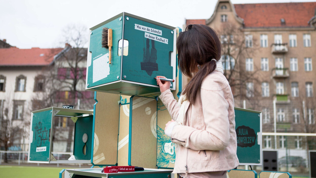 Eine Frau in einer wei&szlig;en Jacke steht neben einem modularen, t&uuml;rkisfarbenen Ausstellungskoffer, der Teil der mobilen Ausstellung des J&uuml;dischen Museums Berlin ist. Die Ausstellungsmodule sind so gestaltet, dass sie flexibel in verschiedenen R&auml;umen aufgebaut werden k&ouml;nnen, vor einer st&auml;dtischen Kulisse mit Altbauten im Hintergrund.