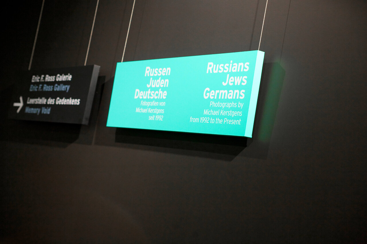 Ausstellungsschild in t&uuml;rkisfarbener Hintergrundbeleuchtung mit dem Text "Russen Juden Deutsche" im J&uuml;dischen Museum Berlin, Teil der Ausstellungsmedien zum Langzeitprojekt von Michael Kerstgens &uuml;ber j&uuml;disches Leben in Deutschland.