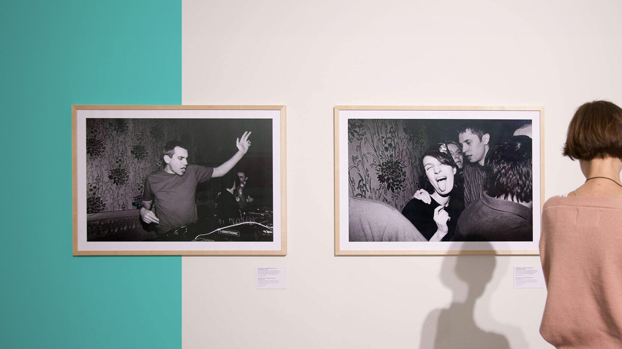 Zwei schwarzwei&szlig;e Fotografien an einer Wand mit t&uuml;rkisfarbenem Hintergrund, eine zeigt eine Person mit gehobener Hand, die andere eine Gruppenaufnahme. Eine Person von hinten betrachtet die Ausstellungsfotos.