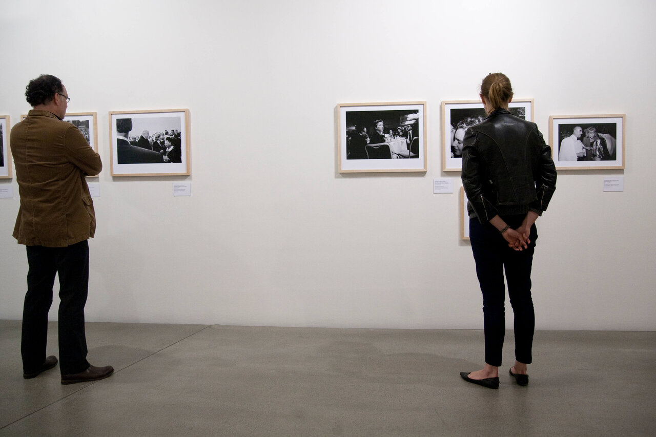 Zwei Personen stehen vor Schwarzwei&szlig;fotografien in einer Galerie, einer in brauner Jacke links und eine in schwarzer Jacke rechts, beide mit dem R&uuml;cken zur Kamera und die Kunstwerke betrachtend.