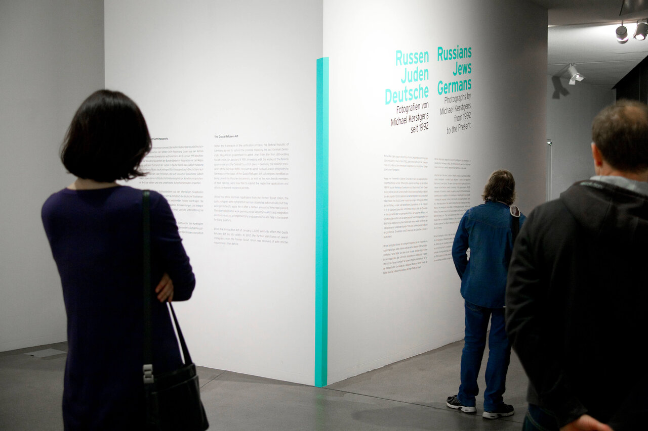 Menschen stehen vor Ausstellungsw&auml;nden mit Texten und einer schmalen t&uuml;rkisfarbenen S&auml;ule in einem Museumsraum, die Besucher sind von hinten zu sehen und betrachten die Ausstellungsinformationen.