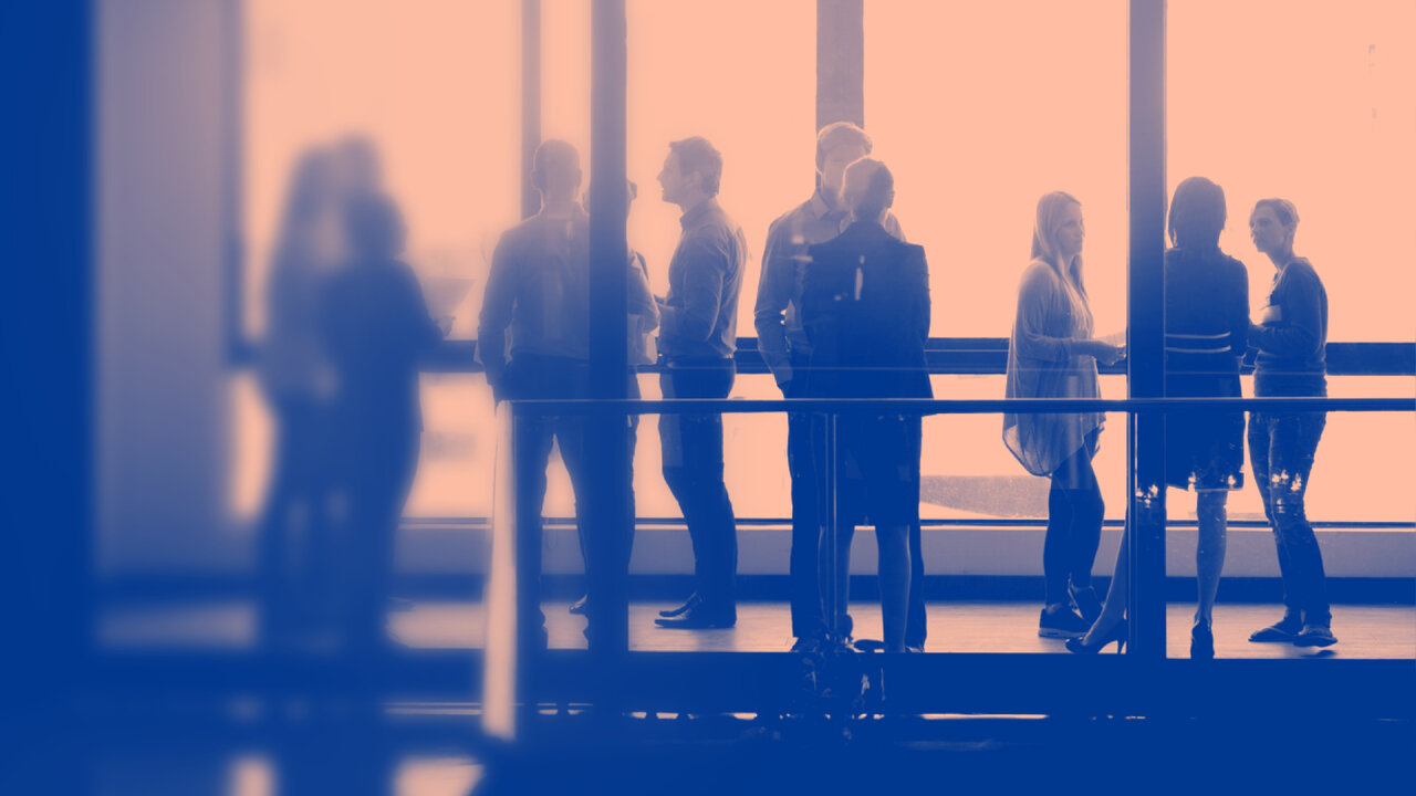 Silhouetten von Personen an einem Gel&auml;nder, verschwommen vor einem warmen, orangefarbenen Hintergrund mit einem blauen Farbverlauf, die eine b&uuml;rom&auml;&szlig;ige oder Konferenz-Atmosph&auml;re suggerieren.