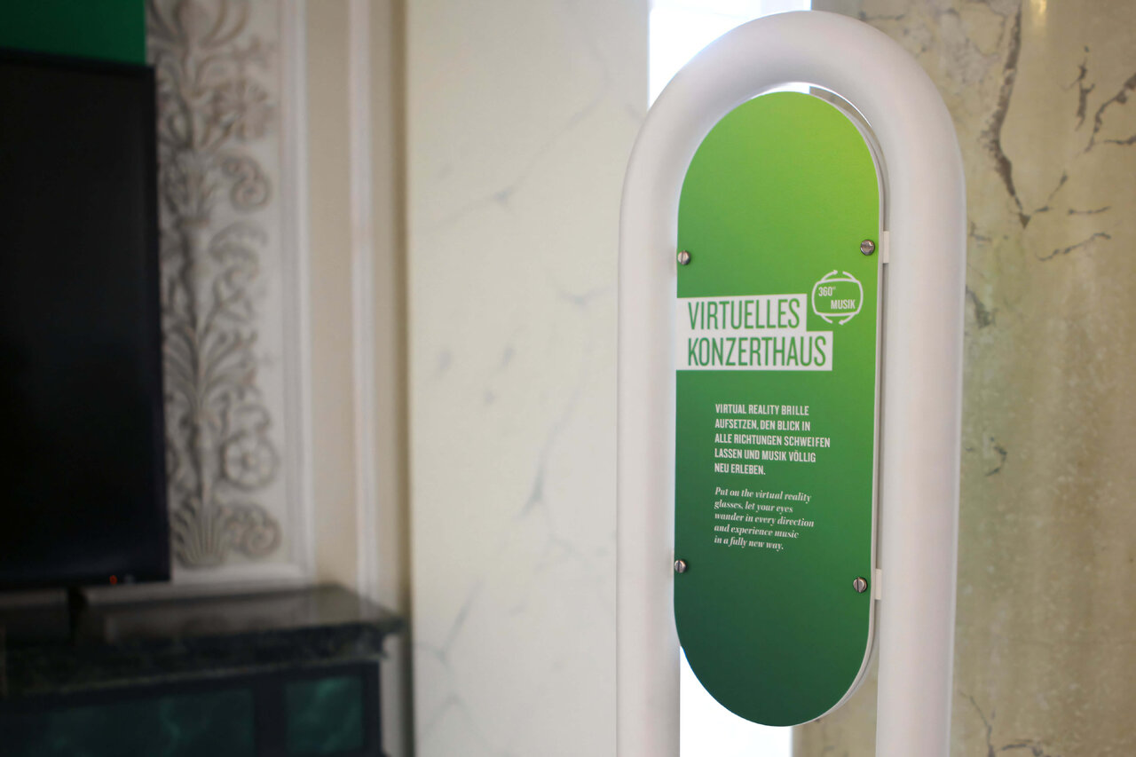 Gr&uuml;ne, ovale Informationstafel mit der Aufschrift "Virtuelles Konzerthaus" an einer wei&szlig;en Wand, die Teil einer kreativen Projektinstallation zur Visualisierung von Mozarts Musik in einer VR-Erfahrung ist.