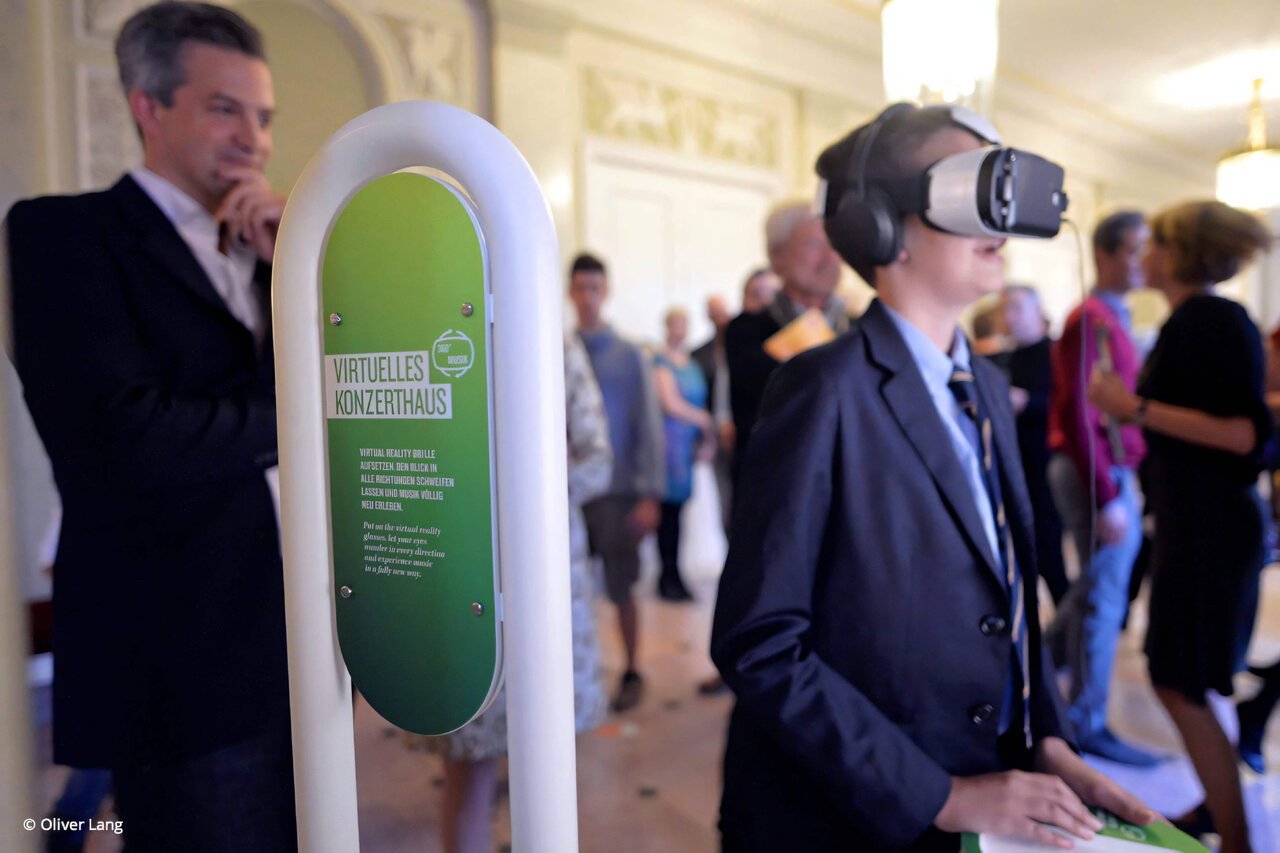 Ein Besucher mit VR-Brille steht neben einer gr&uuml;nen Informationss&auml;ule in einem eleganten Saal, w&auml;hrend im Hintergrund weitere Personen zu sehen sind. Die VR-Installation ist Teil eines innovativen 360&deg;-Konzerterlebnisses von Mozarts Jupiter-Sinfonie, entwickelt in Zusammenarbeit mit dem Konzerthaus Berlin und der HTW.