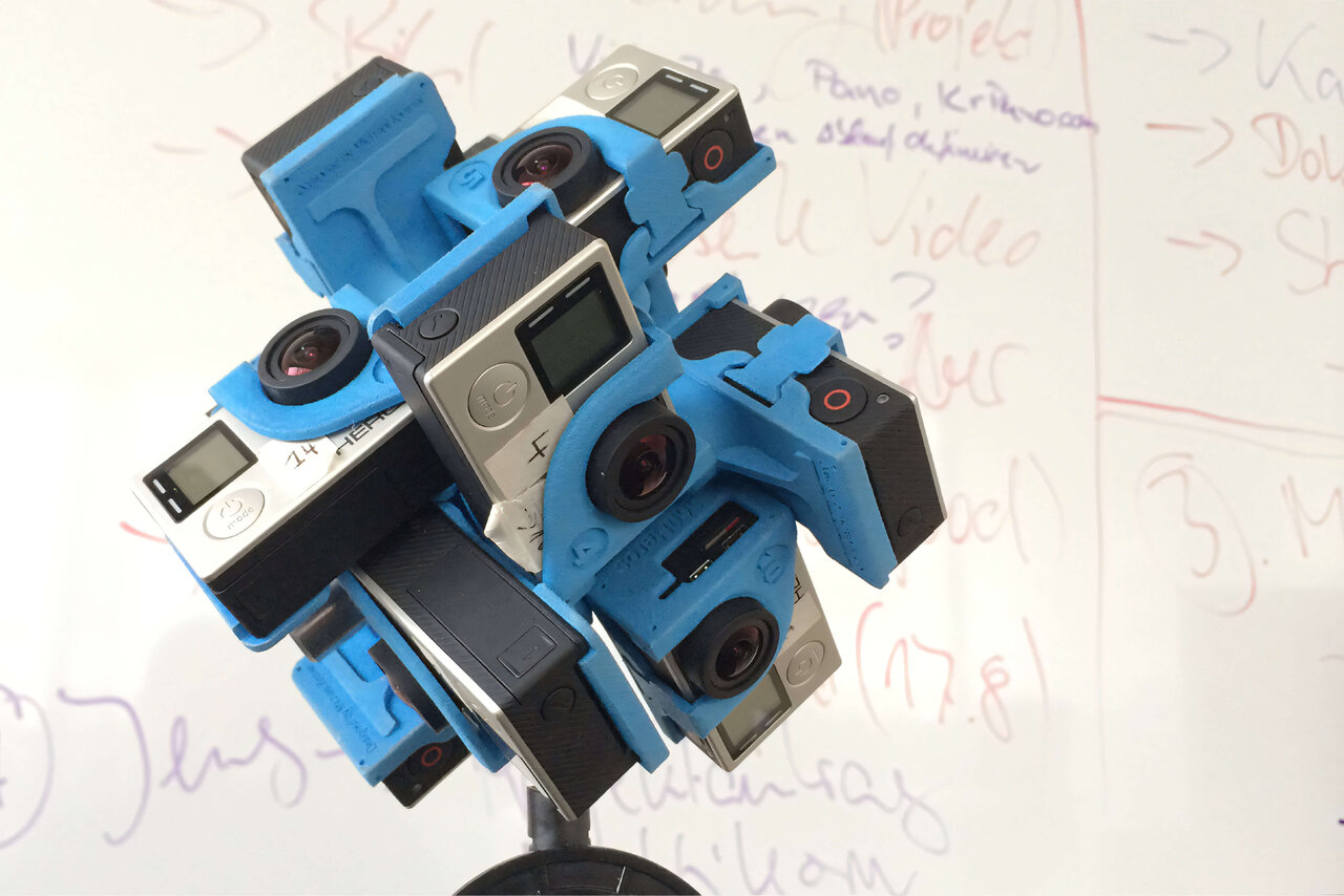Mehrere GoPro-Kameras in blau und grau, so zusammengebaut, dass sie einen 360-Grad-Kameraaufbau f&uuml;r ein innovatives VR-Konzerterlebnis bilden, montiert auf einem Stativ vor einem Hintergrund mit handschriftlichen Notizen.