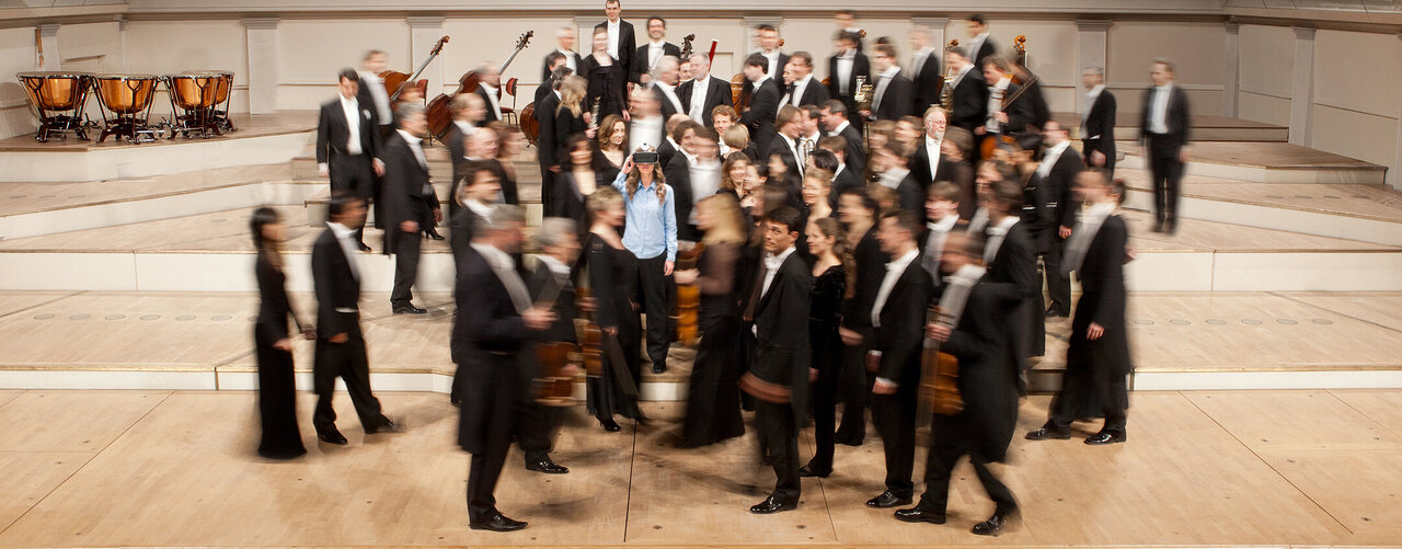 Verschwommenes Gruppenfoto eines Orchesters in Abendgarderobe, die einen Moment der Bewegung und Dynamik einf&auml;ngt, mit Musikinstrumenten und einer leicht zentralen Figur in hellem Hemd.