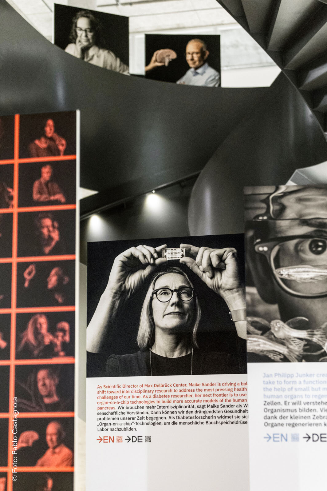 Schwarzwei&szlig;es Portr&auml;t einer Wissenschaftlerin mit Brille, die eine Linse oder ein technisches Ger&auml;t &uuml;ber ihren Kopf h&auml;lt. Im Hintergrund sind weitere Portr&auml;tfotos in verschiedenen Ansichten und Formaten zu sehen, die Teil eines Ausstellungs- oder Projektdesigns zu sein scheinen.
