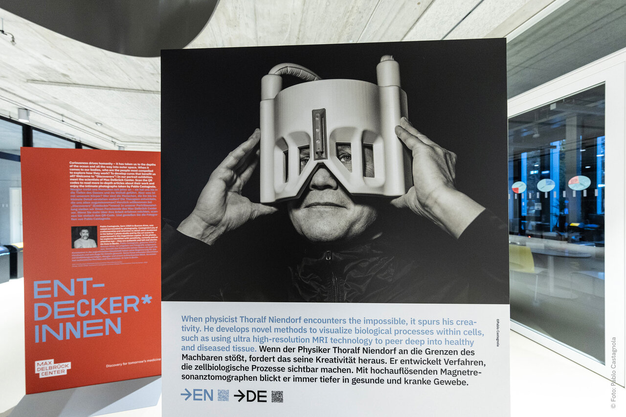 Schwarzwei&szlig;fotografie einer Person, die eine technische Vorrichtung vor den Augen h&auml;lt, die wie eine futuristische Sichtmaschine oder Brille gestaltet ist, in einem modernen Ausstellungsraum mit rotem Informationsschild.