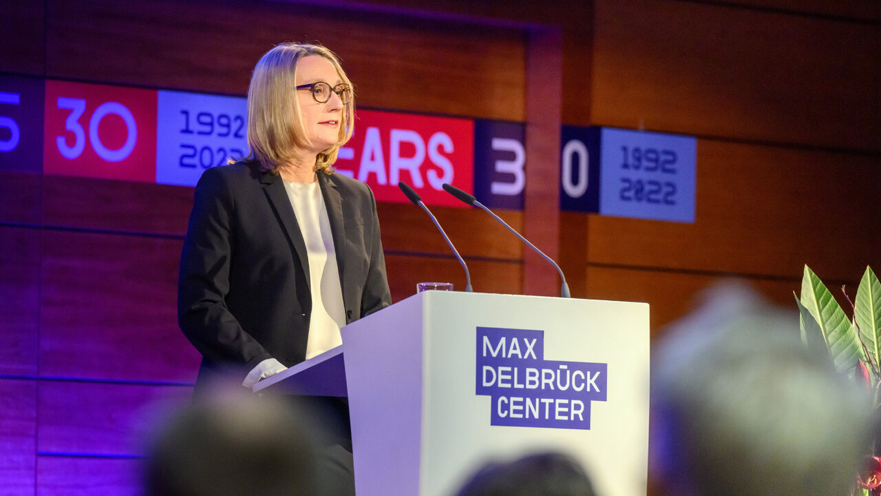 Eine Frau mit Brille und blondem Haar spricht an einem Rednerpult mit dem Schriftzug "Max Delbr&uuml;ck Center" vor einem Hintergrund mit roten und blauen Zahlen.