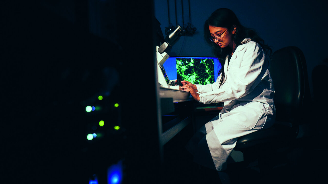 Eine Wissenschaftlerin in wei&szlig;em Labormantel untersucht konzentriert eine leuchtend blaue Petrischale mit gr&uuml;ner Substanz in einem dunklen Labor, umgeben von unscharf leuchtenden gr&uuml;nen und blauen Lichtpunkten.