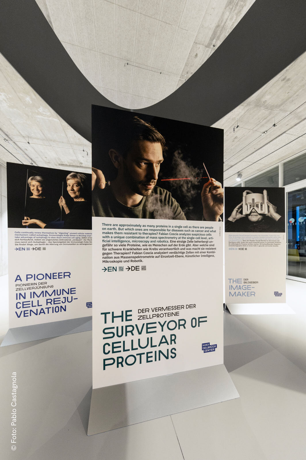 Ausstellungsansicht mit Infotafeln &uuml;ber einen Zellprotein-Forscher, zentral ein gro&szlig;es Portr&auml;tfoto des Wissenschaftlers vor einem minimalistischen, wei&szlig;en Architekturraum mit geometrischen Linien und Kurven.