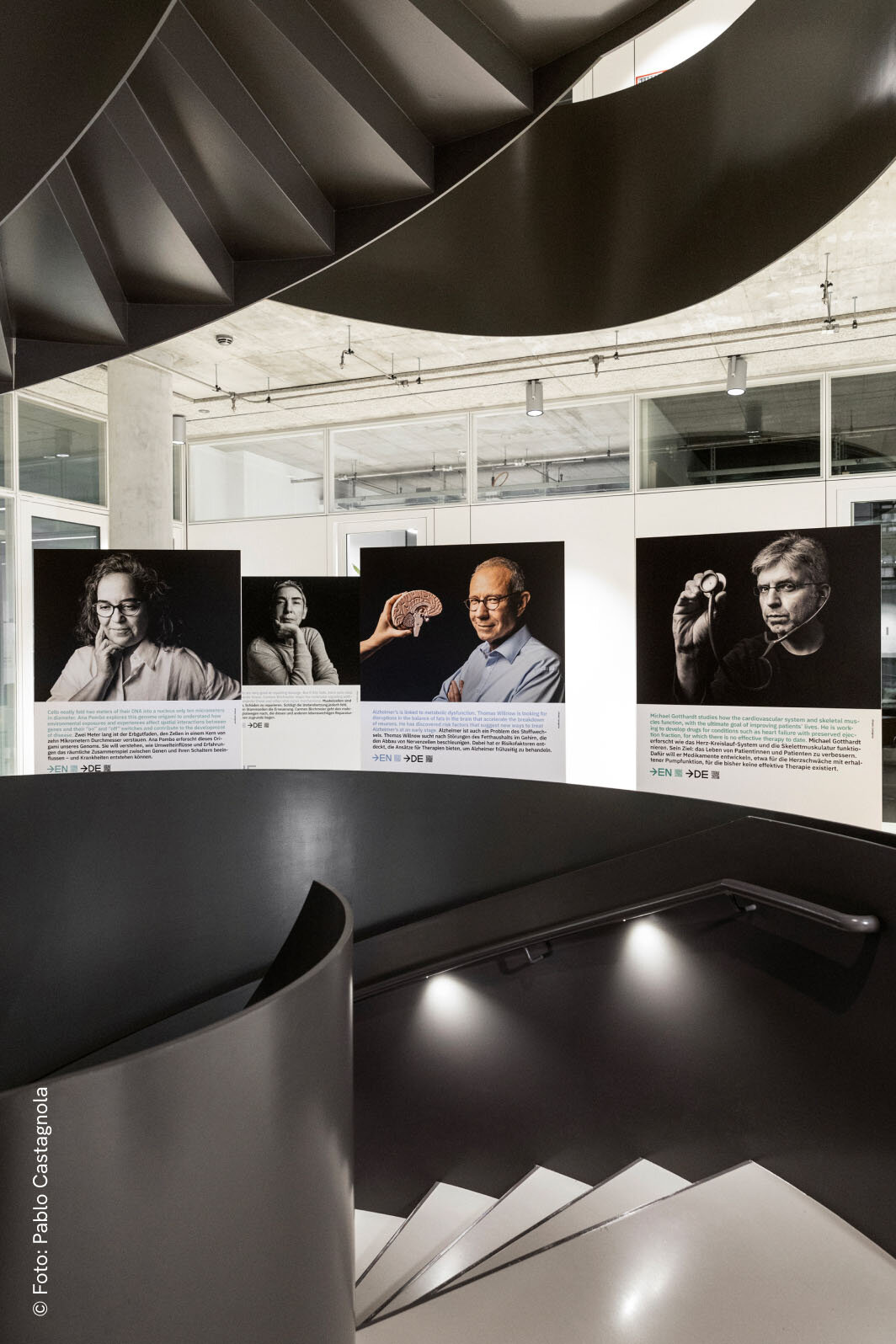 Eine Ausstellungswand mit mehreren Schwarzwei&szlig;portr&auml;ts von Personen in einem modernen, minimalistischen Ausstellungsraum mit geschwungenen schwarzen Elementen und Treppe im Vordergrund.