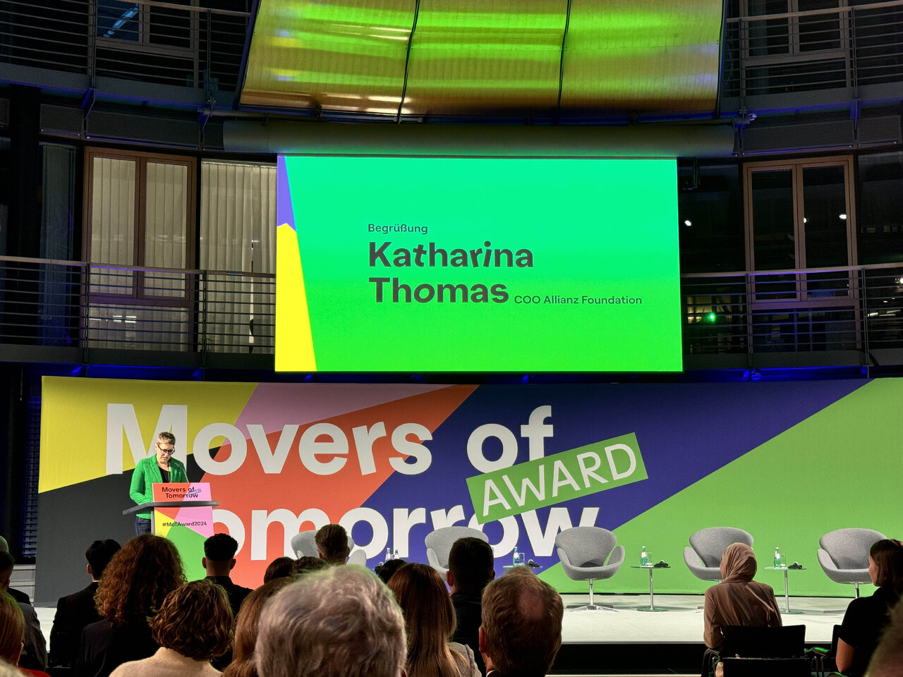Katharina Thomas auf der B&uuml;hne beim "Movers of Tomorrow Award" der Allianz Foundation, mit gr&uuml;n-gelb designtem B&uuml;hnenhintergrund und Publikum im Vordergrund.