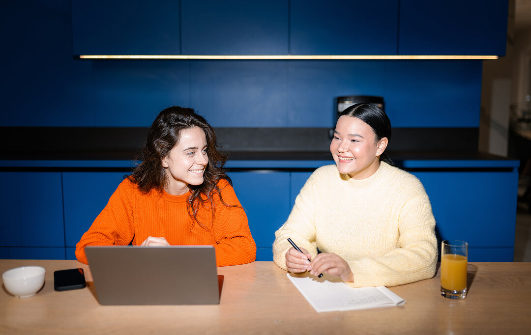 Zwei junge Frauen in orange und gelben Strickpullovern sitzen an einem Tisch mit einem Laptop, l&auml;cheln und arbeiten gemeinsam an einem Projekt.