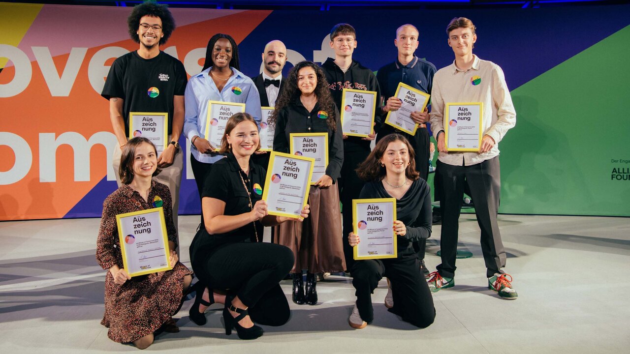 Gruppenfoto von neun Preistr&auml;gern des "Movers of Tomorrow Award" mit Urkunden, die vor einem farbenfrohen Hintergrund stehen und sitzen, gekleidet in verschiedene Alltagsoutfits.
