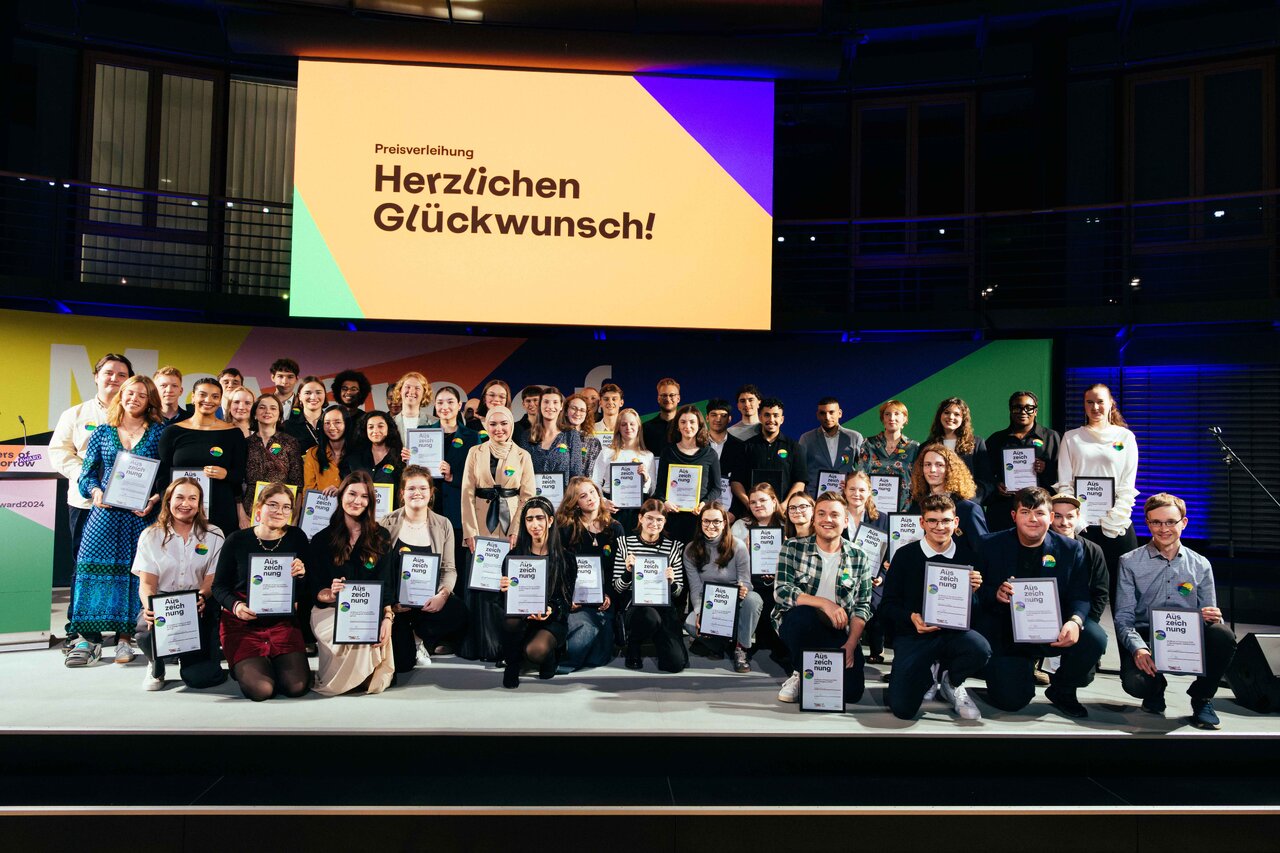 Gruppenfoto der Preistr&auml;ger des "Movers of Tomorrow Award" der Allianz Foundation, die auf einer B&uuml;hne mit einem gro&szlig;en Gratulationsbildschirm im Hintergrund stehen und Urkunden halten.